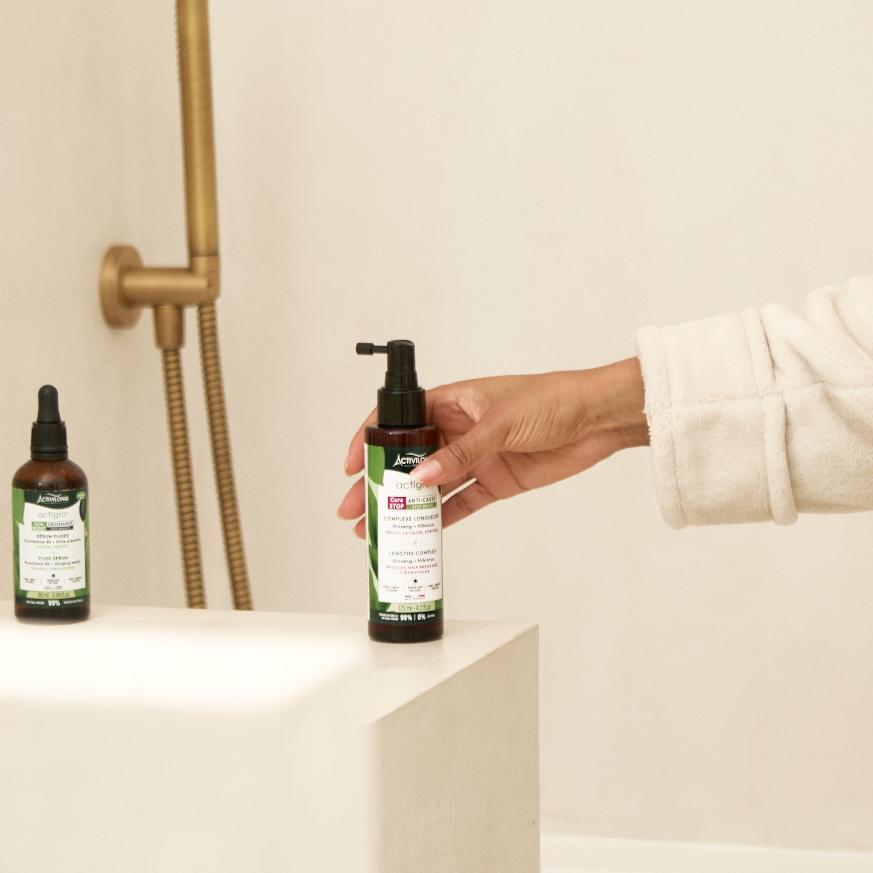Hand holding an ACTIVILONG ACTIGRO Sérum Complexe Longueurs 125ml bottle by ABS France Wholesale with another bottle on a shelf in a bathroom setting