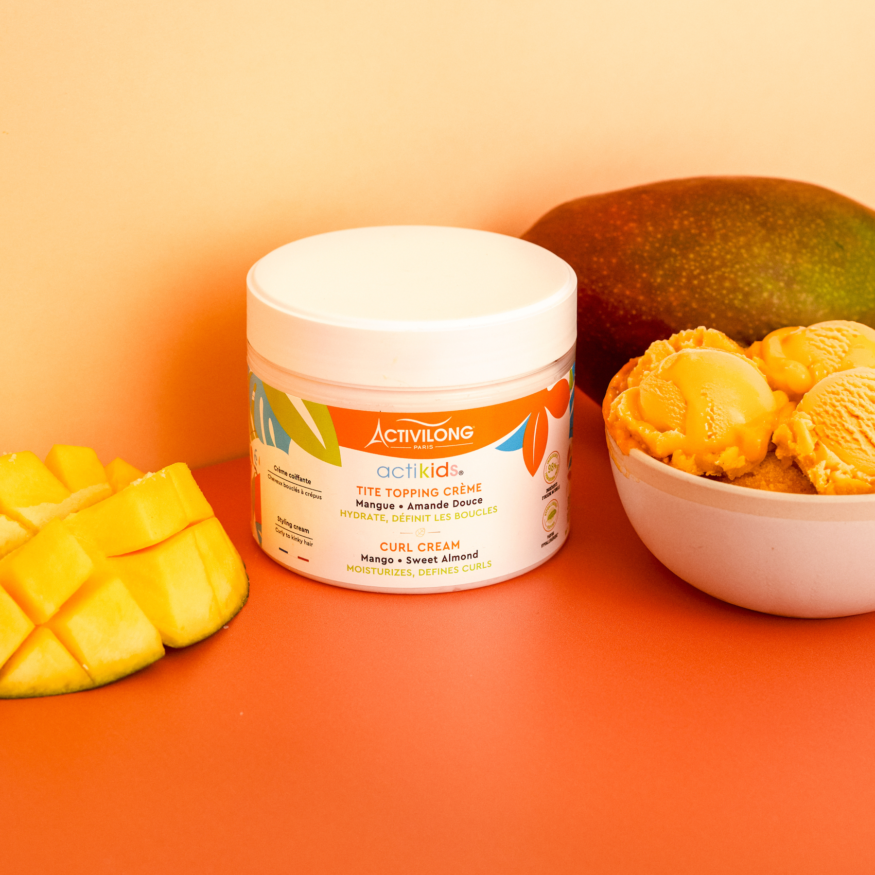 Jar of ACTIVILONG ACTIKIDS Tite Topping Crème 300ml with mango slices and a bowl of cookies on an orange background