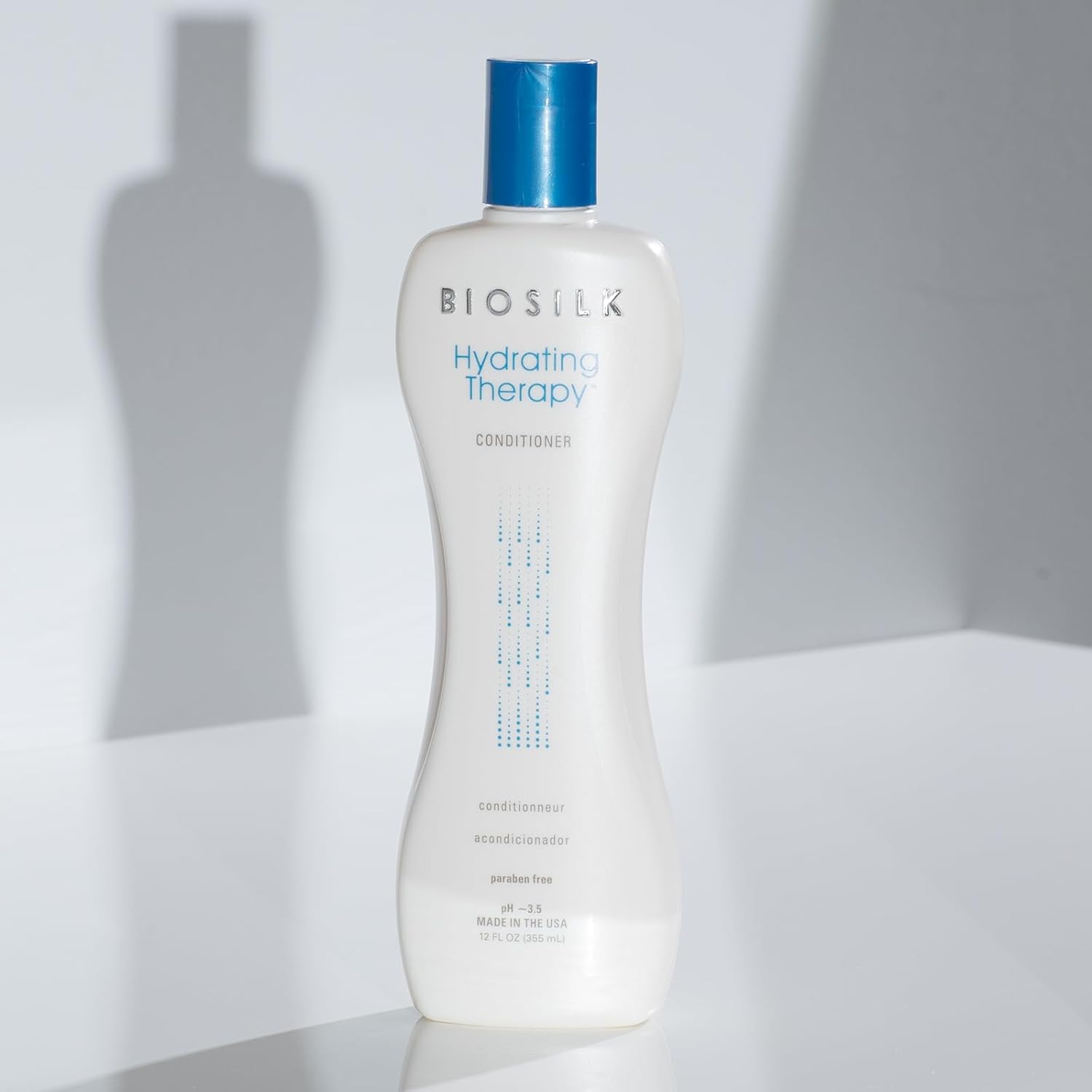 A white bottle of BIOSILK Hydrating Therapy Conditioner with a blue cap, standing on a white surface with a soft shadow cast on the wall behind it.
