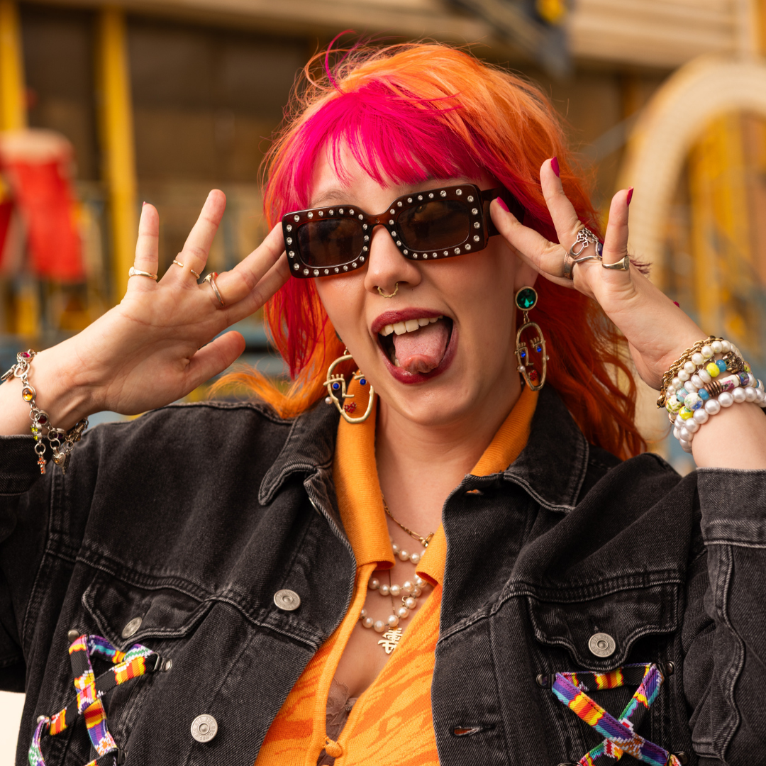 Crazy Color's Person with colorful hair and sunglasses making a peace sign, wearing an orange shirt and black jacket - ABS France Wholesale