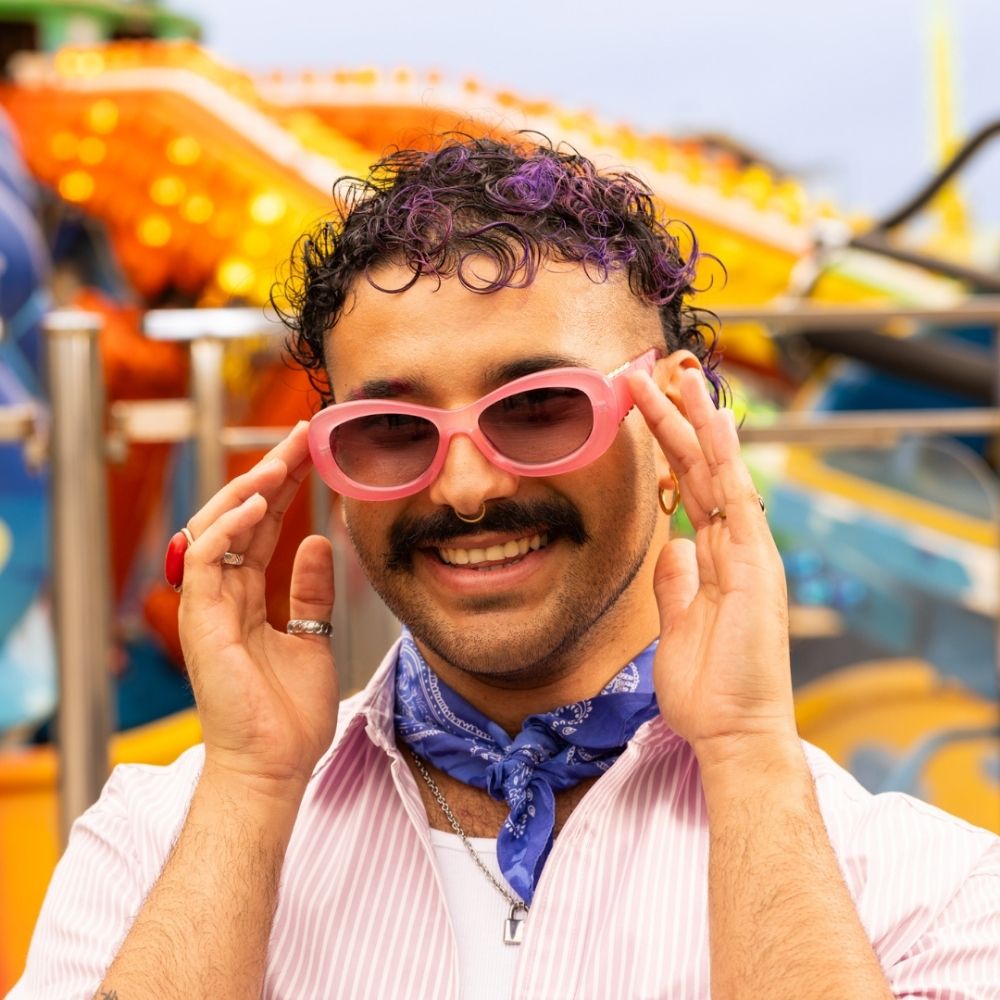 Crazy Color's person wearing pink sunglasses and a blue bandana, with colorful fairground rides in the background by ABS France Wholesale