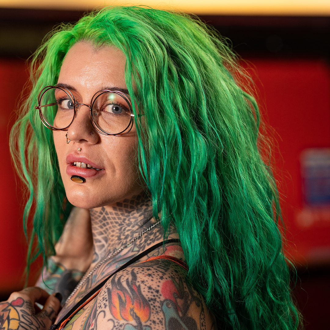 Crazy Color's Person with green hair and tattoos wearing glasses, against a blurred red background by ABS France Wholesale
