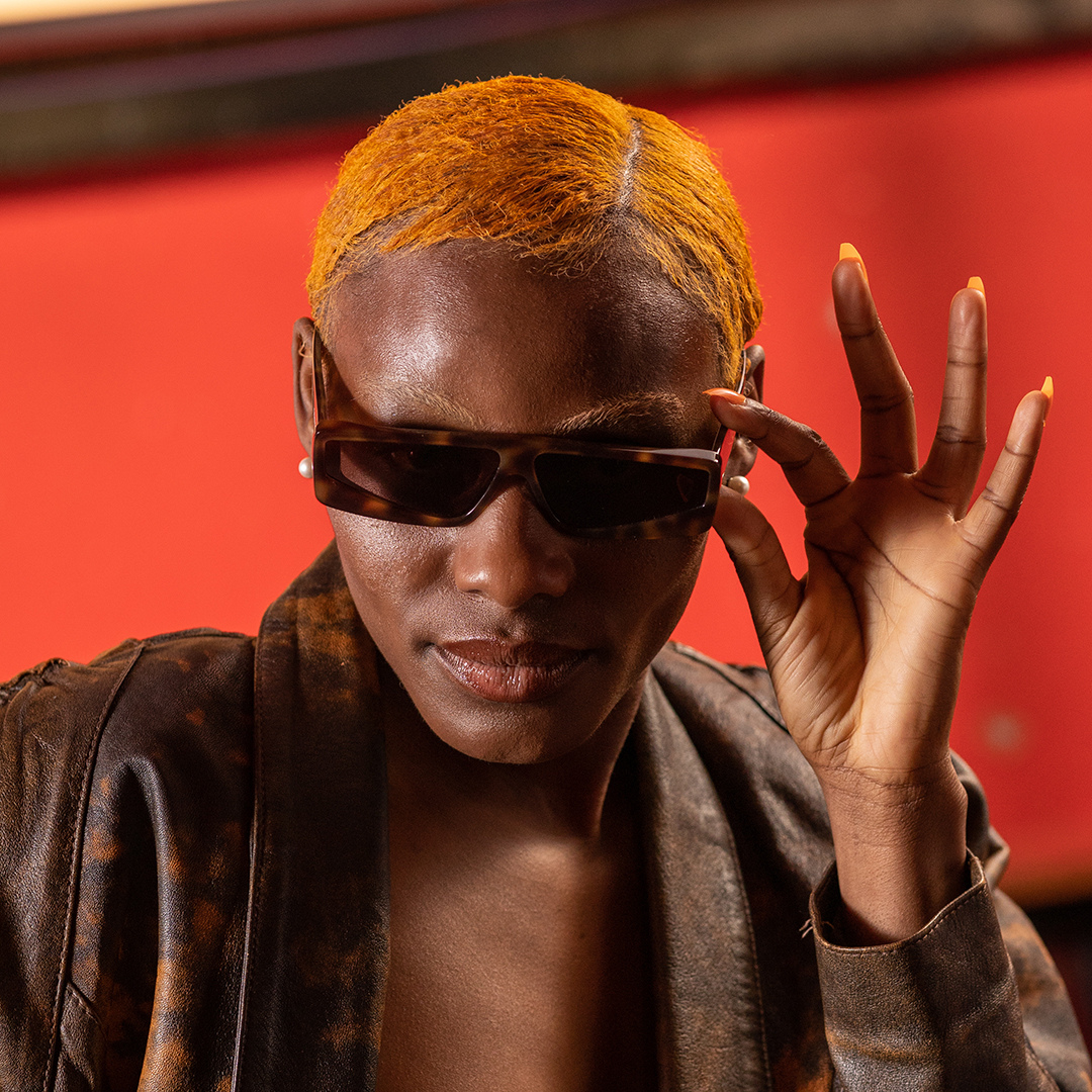 Crazy Color's Person with orange hair wearing sunglasses and a brown jacket against a red background by ABS France Wholesale