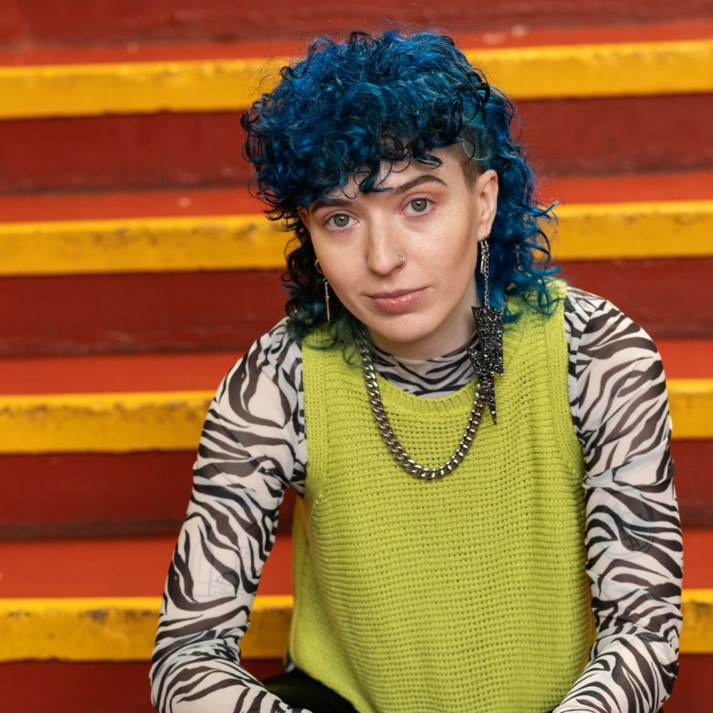 Crazy Color's Person with blue hair and a green sweater sitting on colorful steps by ABS France Wholesale