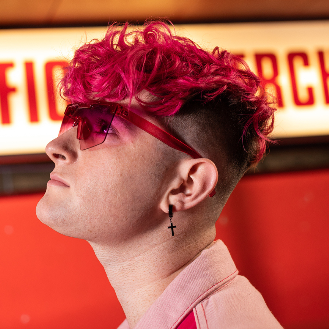Crazy Color's Person with pink hair and sunglasses looking to the side, against a red and yellow background by ABS France Wholesale