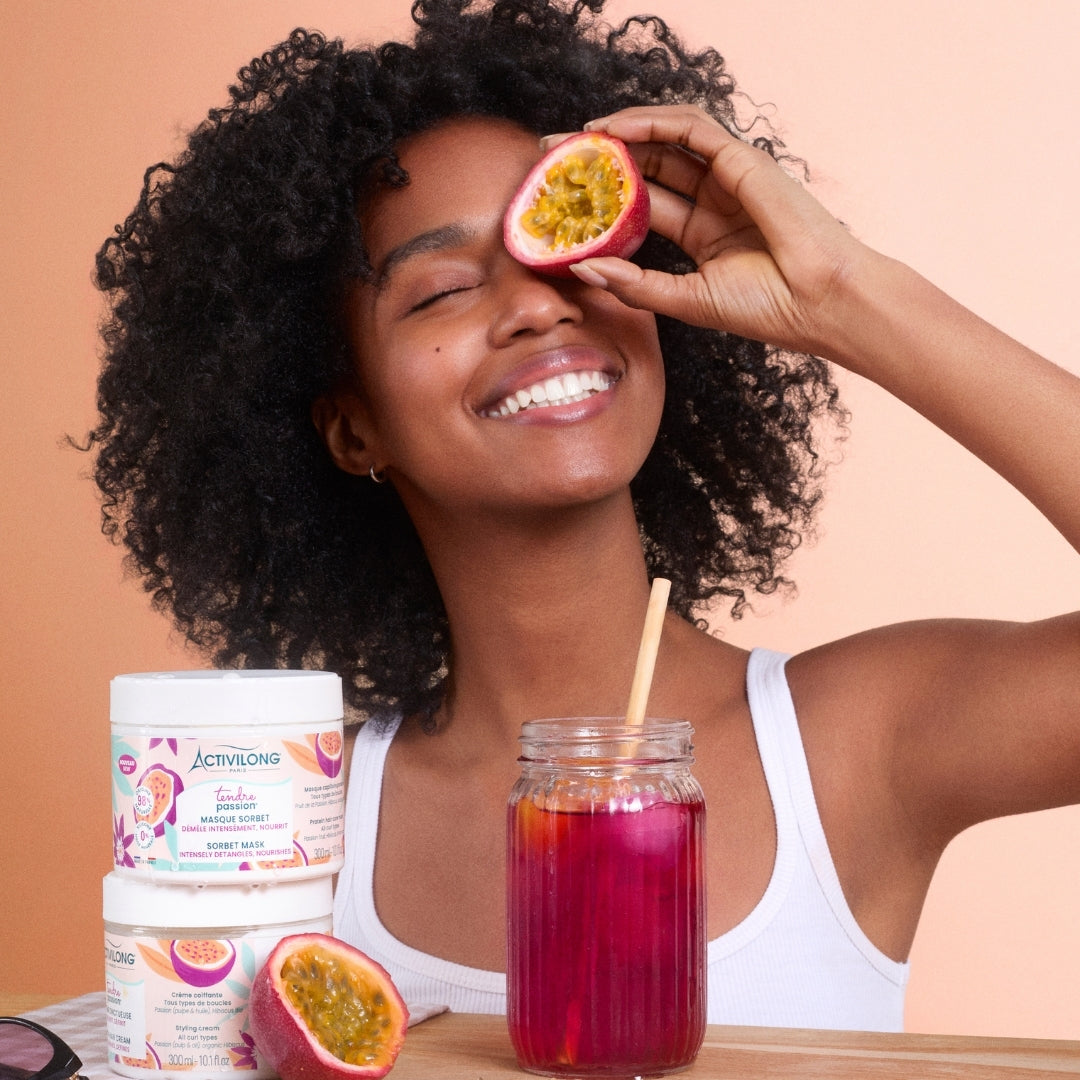 Woman holding a passion fruit half, with ACTIVILONG TENDRE PASSION Masque Sorbet 300ml jars by ABS France Wholesale and a drink on a table.