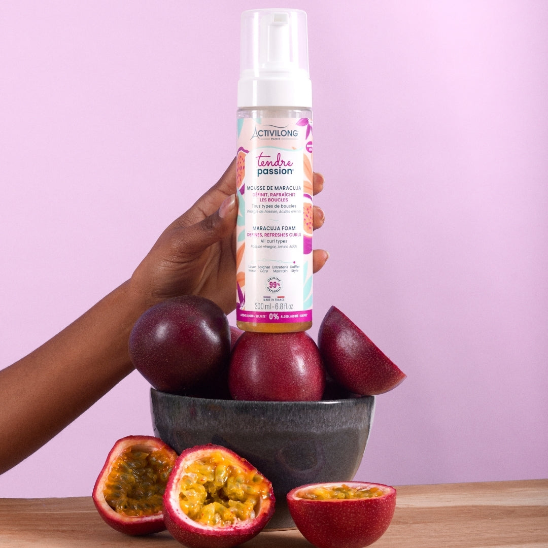 Hand holding an ACTIVILONG TENDRE PASSION Mousse de Maracuja 200ml foam bottle by ABS France Wholesale with passion fruits on a pink background