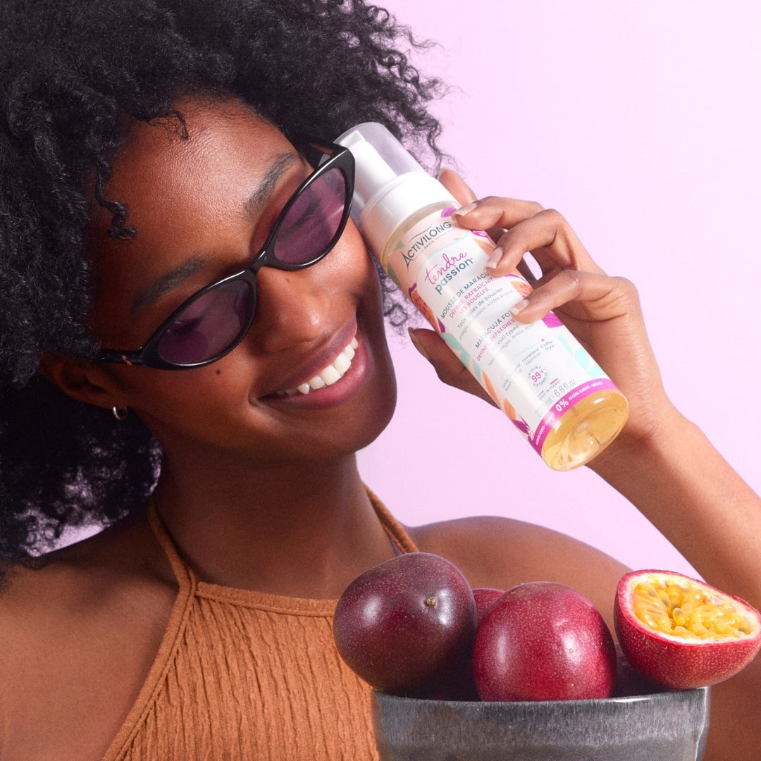 Woman holding an ACTIVILONG TENDRE PASSION Mousse de Maracuja 200ml foam bottle by ABS France Wholesale with fruits on a pink background
