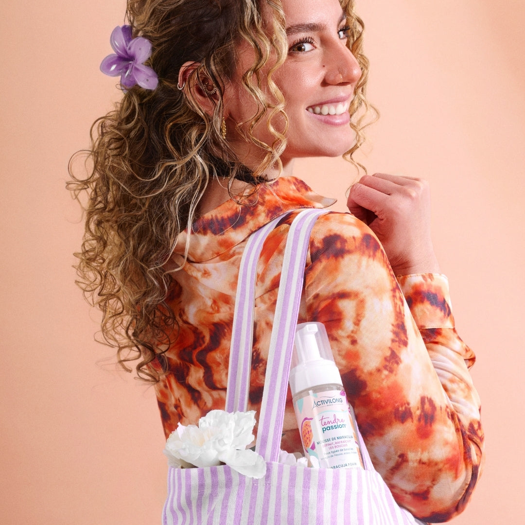 Woman with floral hair accessory holding a tote bag with an ACTIVILONG TENDRE PASSION Mousse de Maracuja 200ml foam bottle by ABS France Wholesale on a peach background