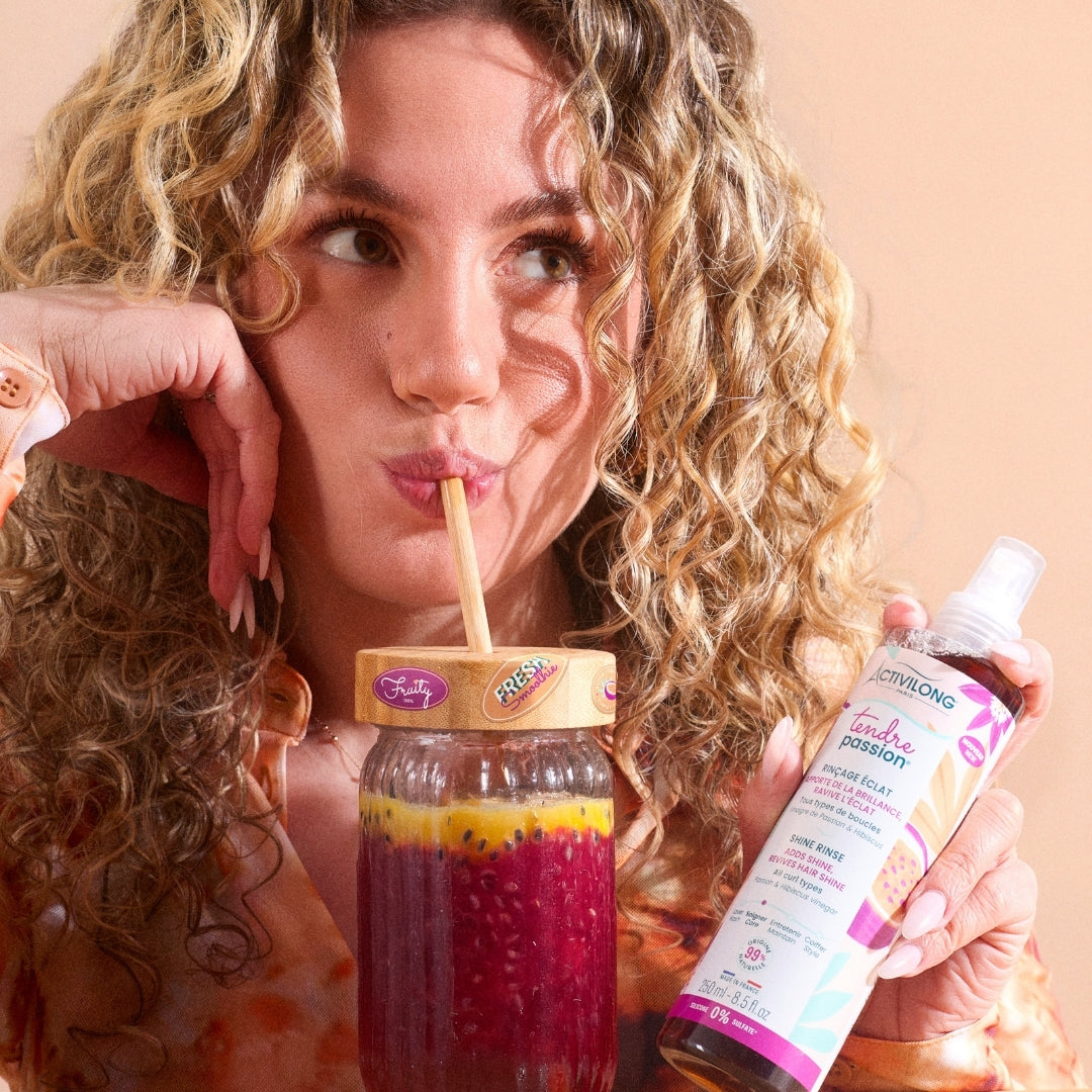 Woman drinking a colorful juice from a glass with a straw, holding an ACTIVILONG TENDRE PASSION Rinçage Éclat 250ml bottle by ABS France Wholesale against a peach background.
