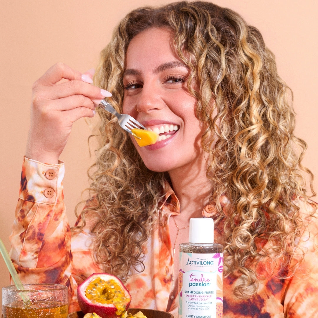 Woman eating passion fruit with a bottle of ACTIVILONG TENDRE PASSION Shampooing Fruité 250ml by ABS France Wholesale in the foreground