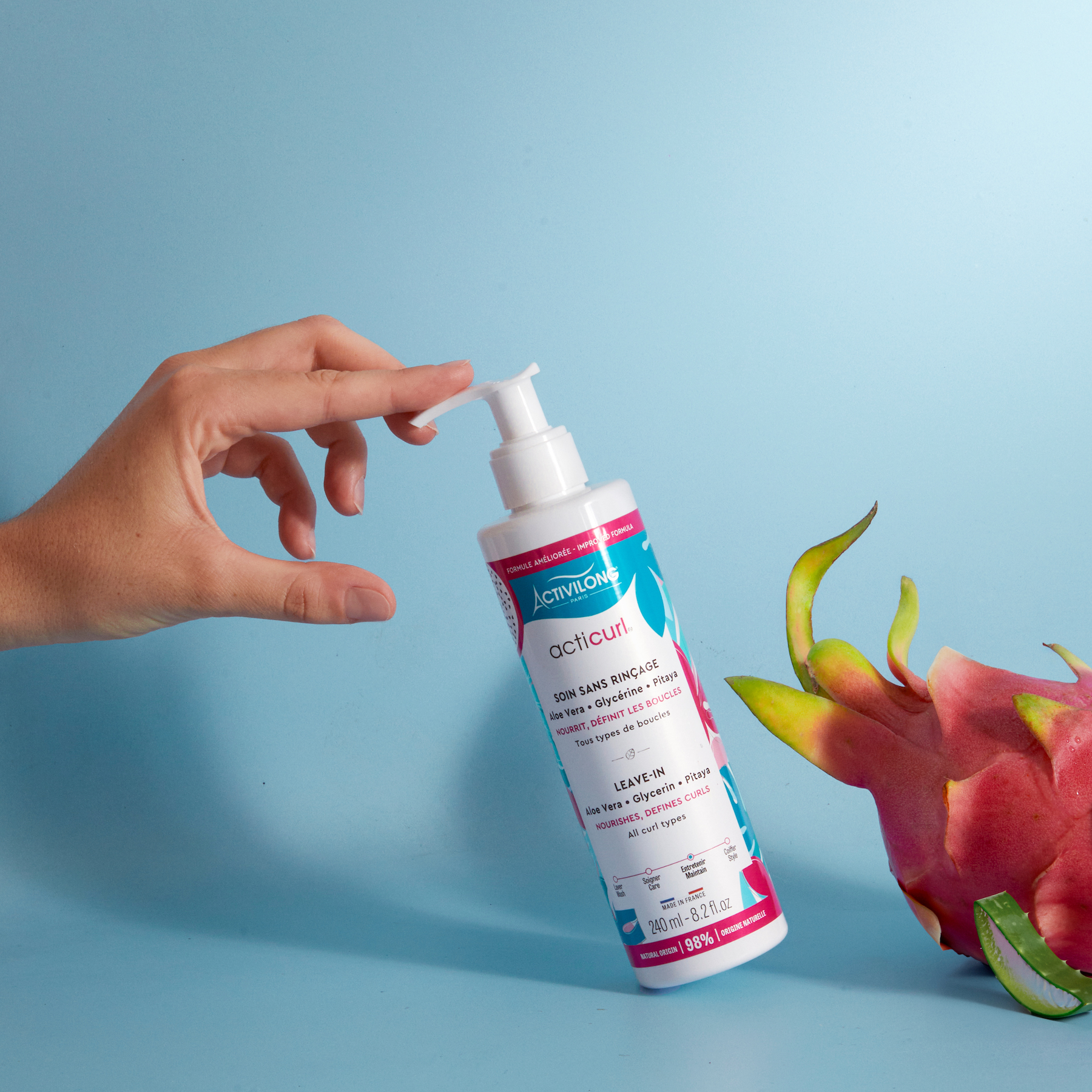 Hand holding a bottle of ACTIVILONG ACTICURL Leave-In 240ml by ABS France Wholesale with a dragon fruit on a blue background