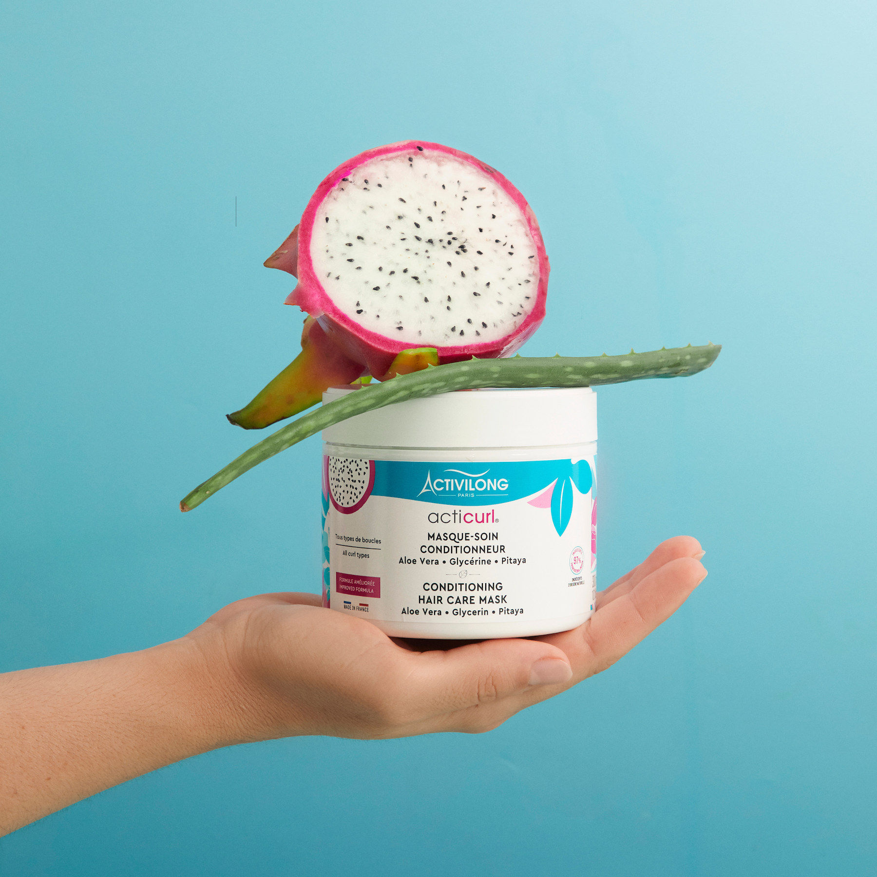 Hand holding a ACTIVILONG ACTICURL Masque 300ml jar by ABS France Wholesale with a dragon fruit on a blue background