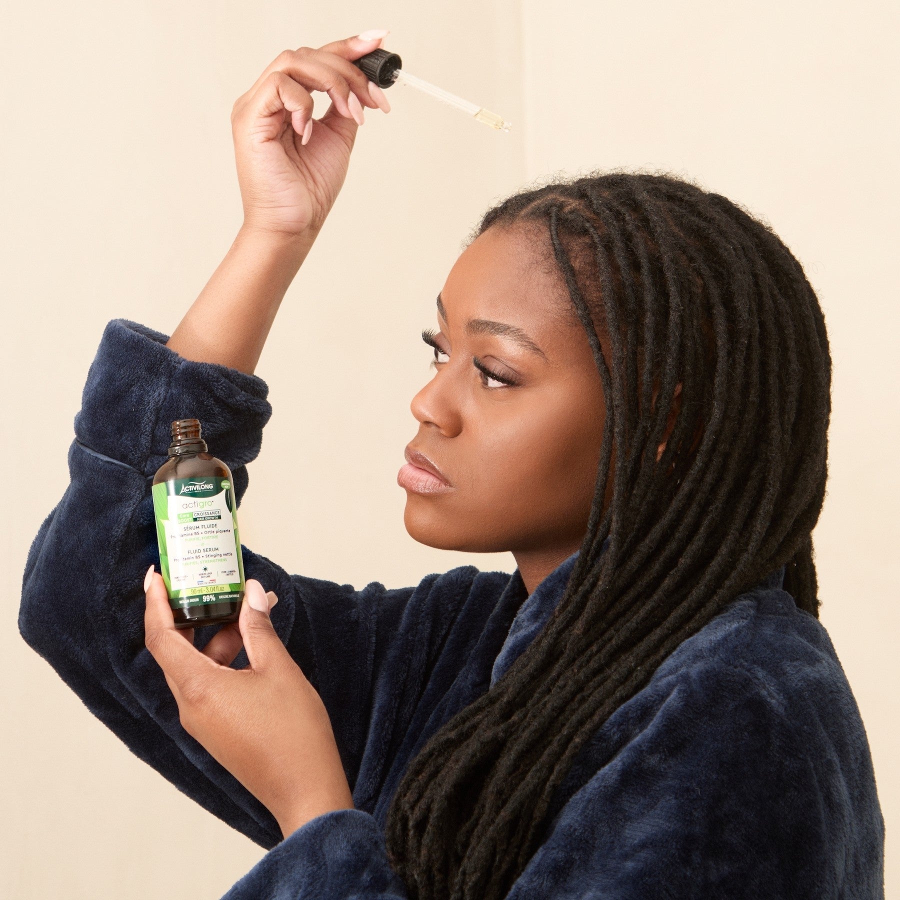 Woman applying a dropper of a Bottle of ACTIVILONG ACTIGRO Sérum Fluide 90ml by ABS France Wholesale contents to her skin against a beige background