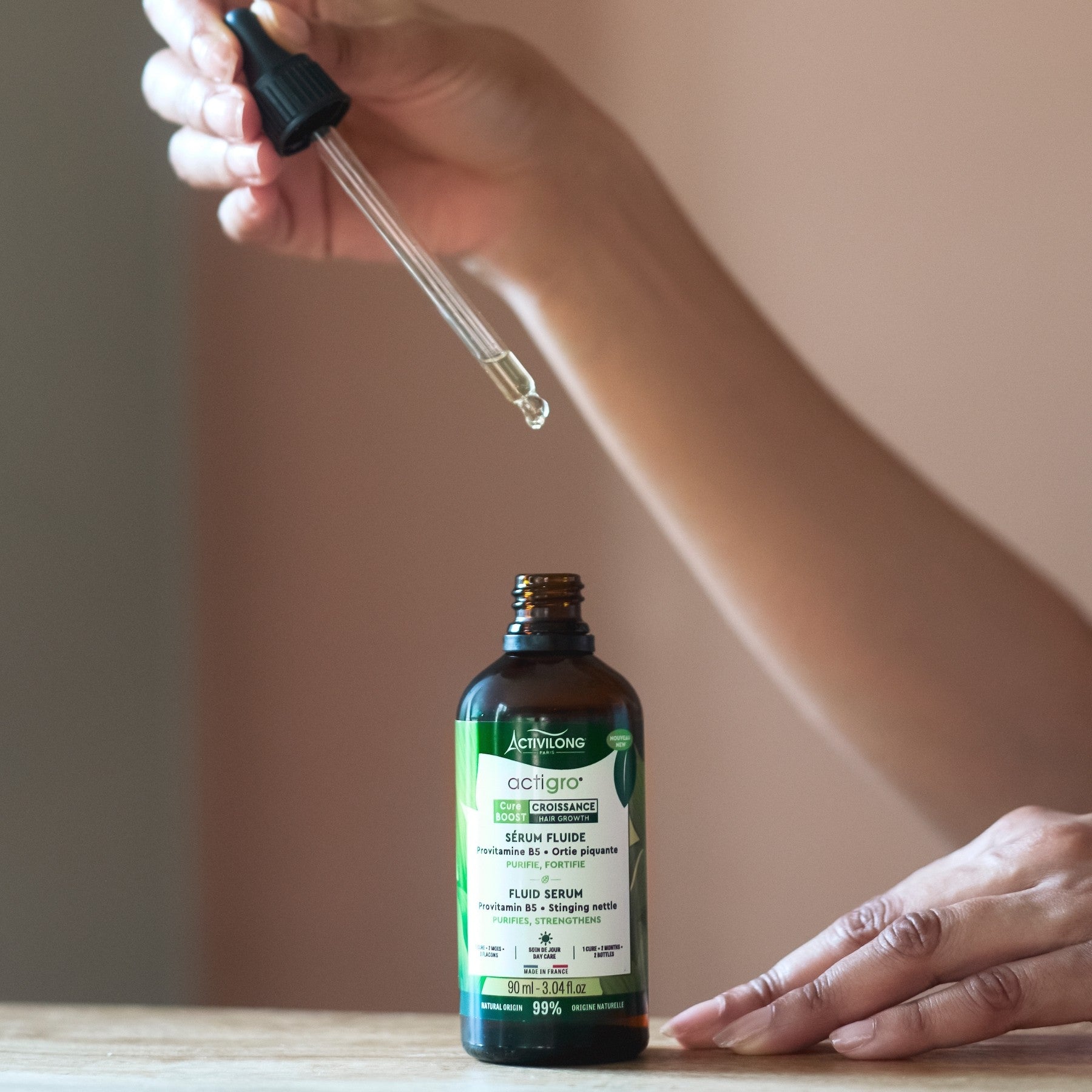 Hand holding a dropper over a Bottle of ACTIVILONG ACTIGRO Sérum Fluide 90ml by ABS France Wholesale on a wooden surface.