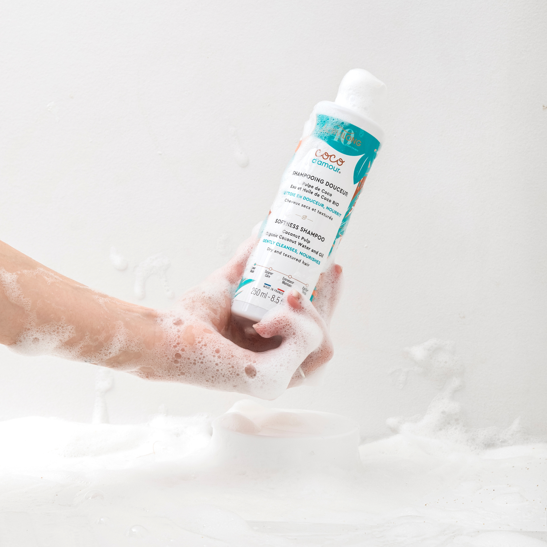 Hand holding a Bottle of ACTIVILONG COCO D’AMOUR Shampooing Douceur 250ml by ABS France Wholesale with a foamy background