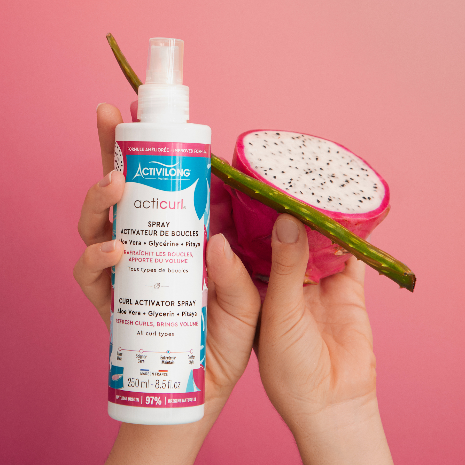 Hand holding a Bottle of ACTIVILONG ACTICURL Spray Activateur 250ml by ABS France Wholesale next to a dragon fruit and aloe vera against a pink background